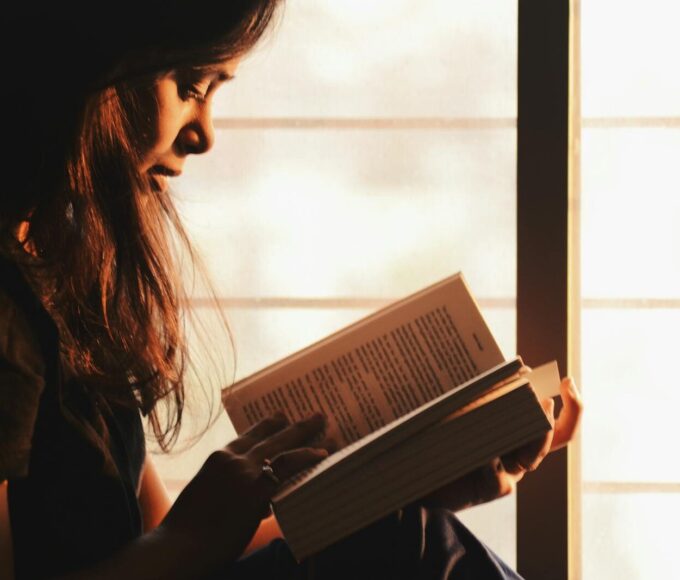 A woman engrossed in reading a book by a sunlit window creates a serene atmosphere.