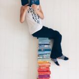 Person casually reading a book while sitting atop a colorful stack of books indoors.