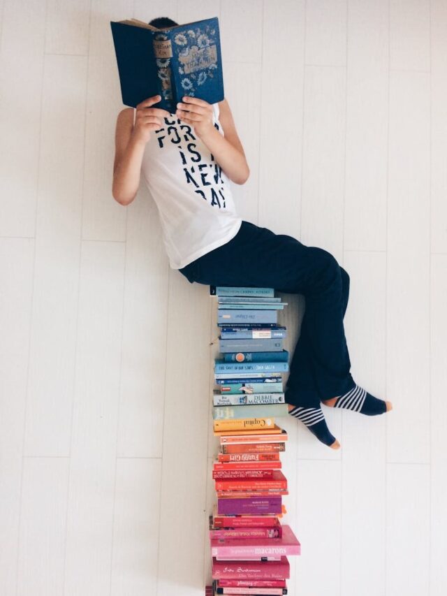 Person casually reading a book while sitting atop a colorful stack of books indoors.