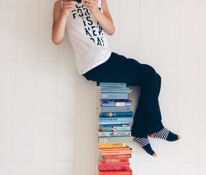 Person casually reading a book while sitting atop a colorful stack of books indoors.