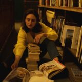 A young woman sits on the floor of a library surrounded by stacks of books, deeply engrossed in reading.