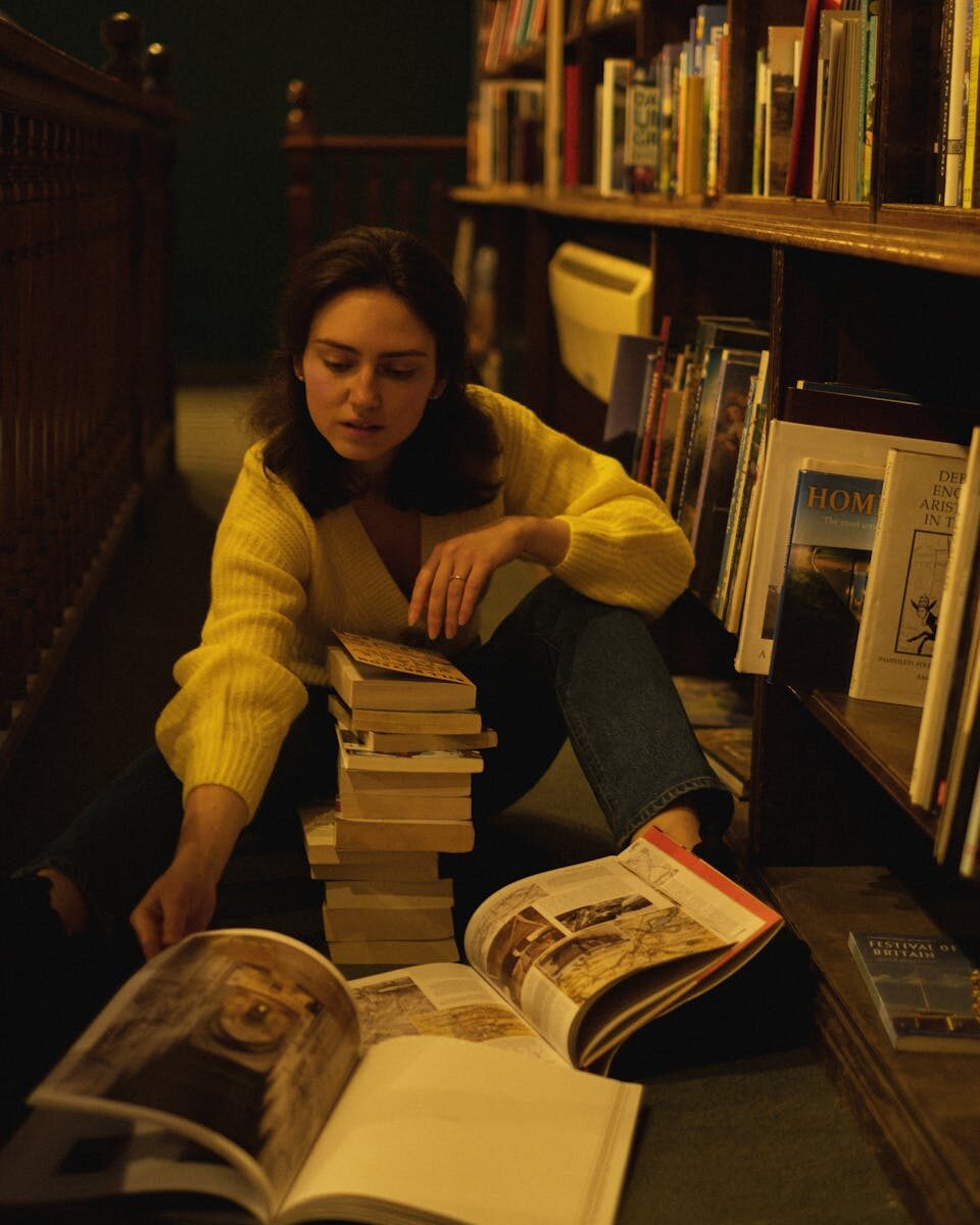 A young woman sits on the floor of a library surrounded by stacks of books, deeply engrossed in reading.