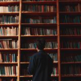 A man with short black hair stands in front of bookshelves in a quiet library.