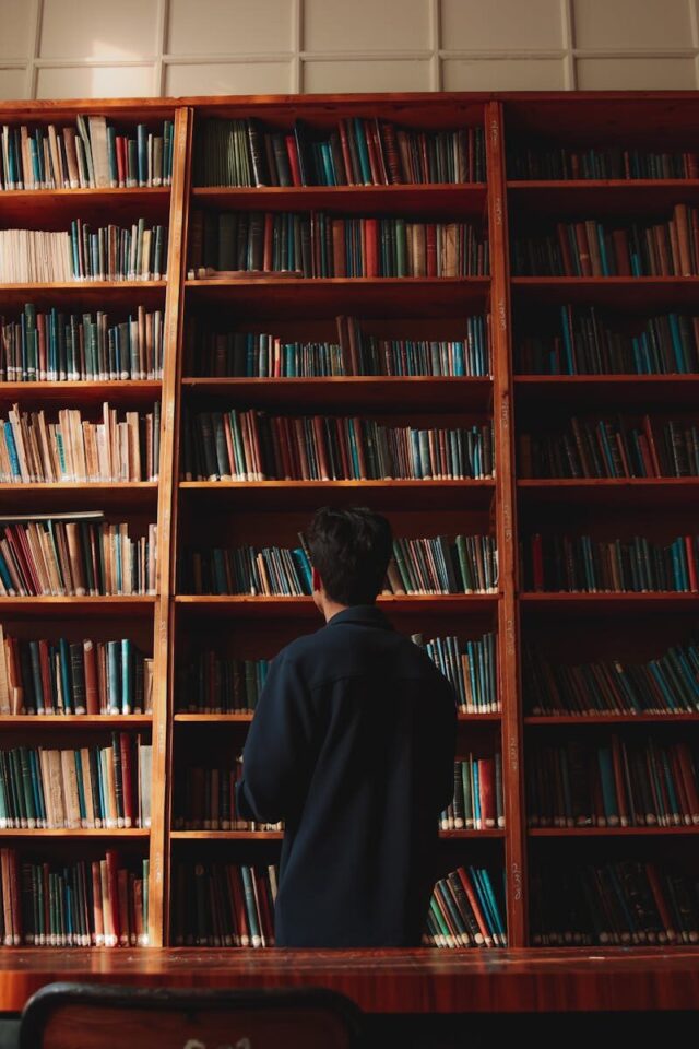 A man with short black hair stands in front of bookshelves in a quiet library.