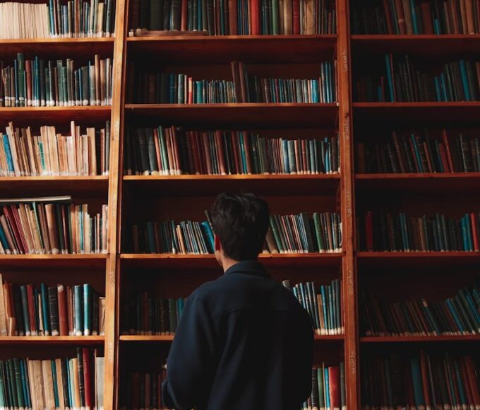 A man with short black hair stands in front of bookshelves in a quiet library.