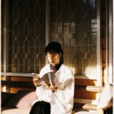 Stylish Asian woman reading a book indoors, bathed in warm sunlight on a cozy bench.