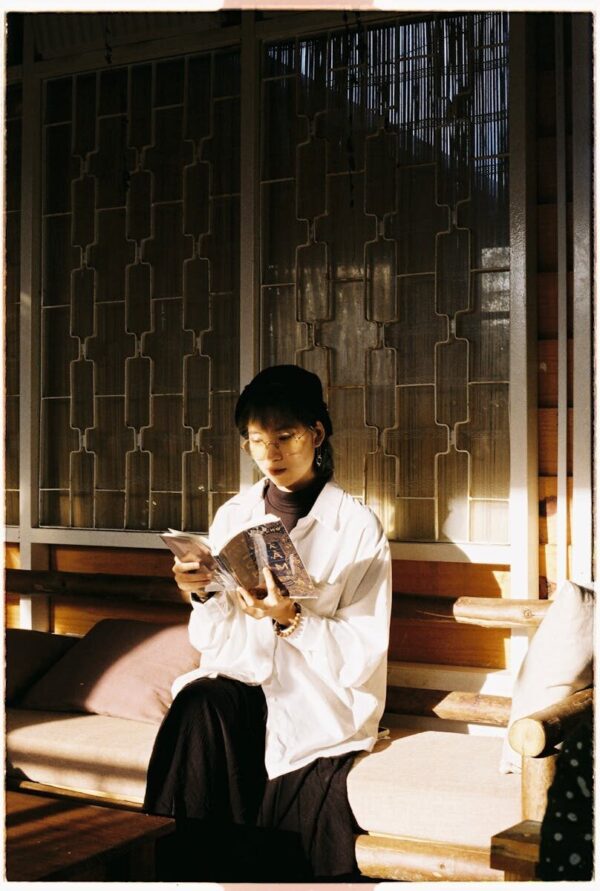 Stylish Asian woman reading a book indoors, bathed in warm sunlight on a cozy bench.