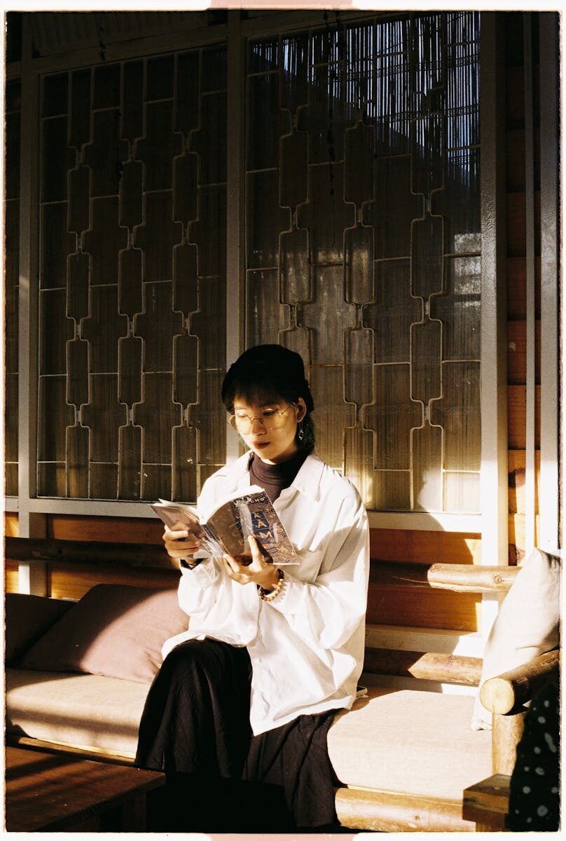Stylish Asian woman reading a book indoors, bathed in warm sunlight on a cozy bench.