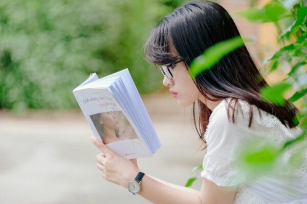 A young woman sits outdoors, absorbed in reading a book during a sunny summer day.