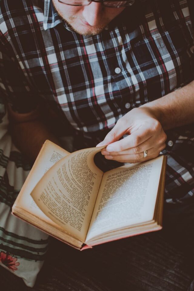 Adult male wearing plaid shirt reading a book indoors for leisure and relaxation.