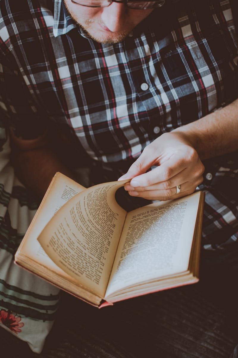 Adult male wearing plaid shirt reading a book indoors for leisure and relaxation.