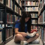 A young woman sitting on the floor of a library, reading a book intently.