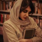 Portrait of a woman in a library, wearing a hijab, holding a book close in a contemplative pose.