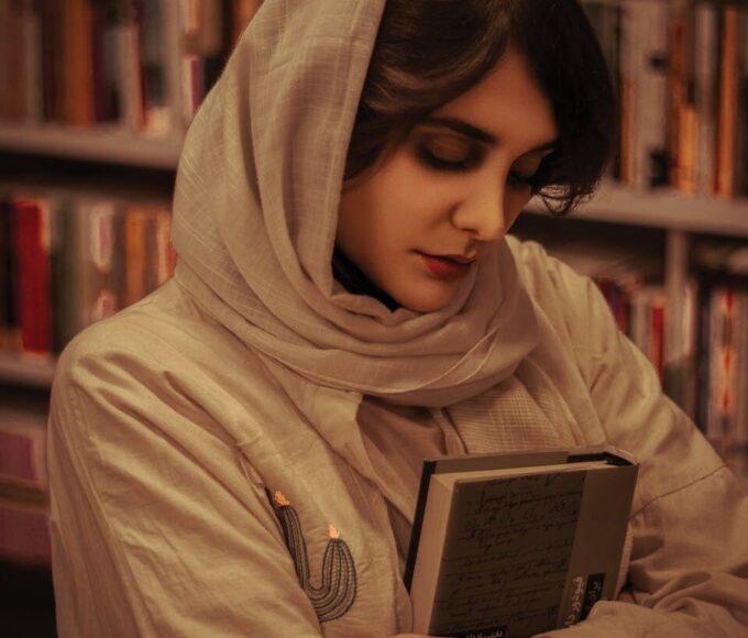 Portrait of a woman in a library, wearing a hijab, holding a book close in a contemplative pose.