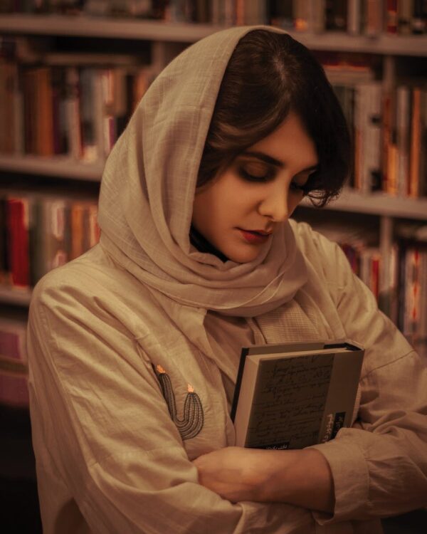 Portrait of a woman in a library, wearing a hijab, holding a book close in a contemplative pose.
