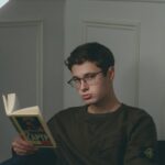 A young man wearing glasses reading a book in a cozy indoor setting at night.