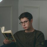 A young man wearing glasses reading a book in a cozy indoor setting at night.