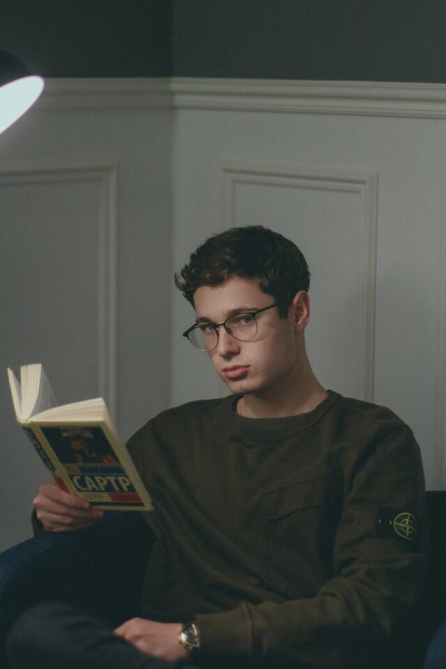 A young man wearing glasses reading a book in a cozy indoor setting at night.