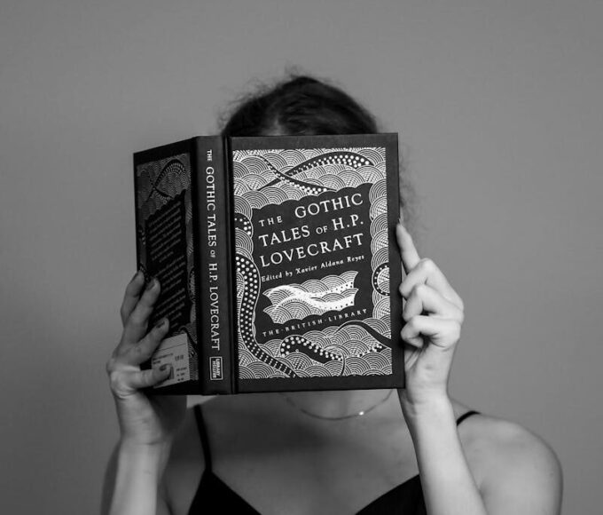 Black and white portrait of a woman reading 'The Gothic Tales of H.P. Lovecraft' in a studio setting.