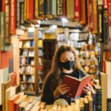 A woman in a mask reading a book in a bookstore surrounded by shelves.