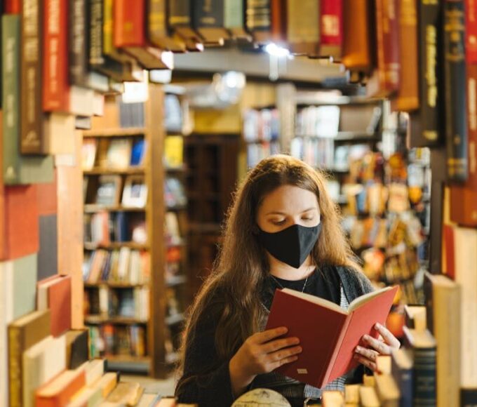 A woman in a mask reading a book in a bookstore surrounded by shelves.