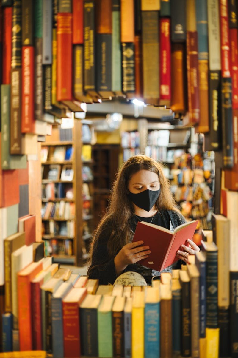 A woman in a mask reading a book in a bookstore surrounded by shelves.