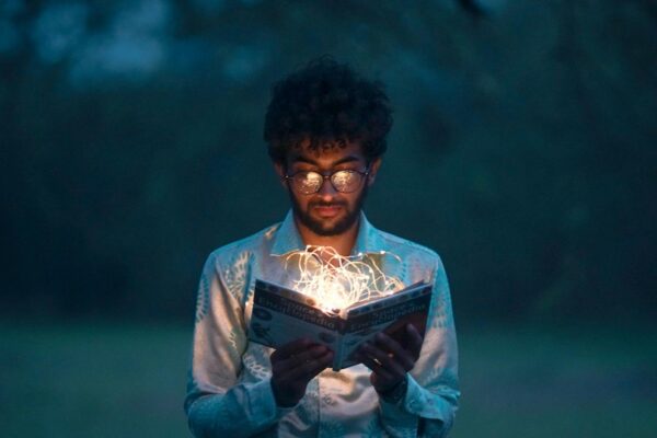 A man with glasses reads an illuminated book outdoors during twilight.