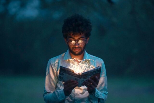 A man with glasses reads an illuminated book outdoors during twilight.