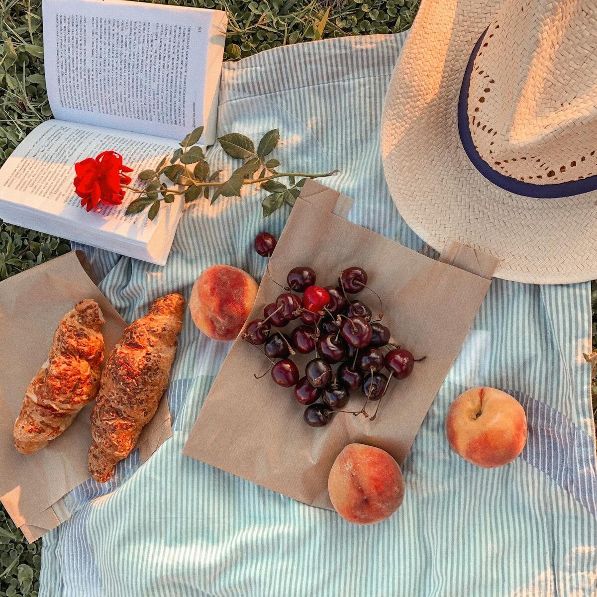 A picturesque summer picnic featuring croissants, cherries, peaches, a hat, and an open book on a striped blanket.