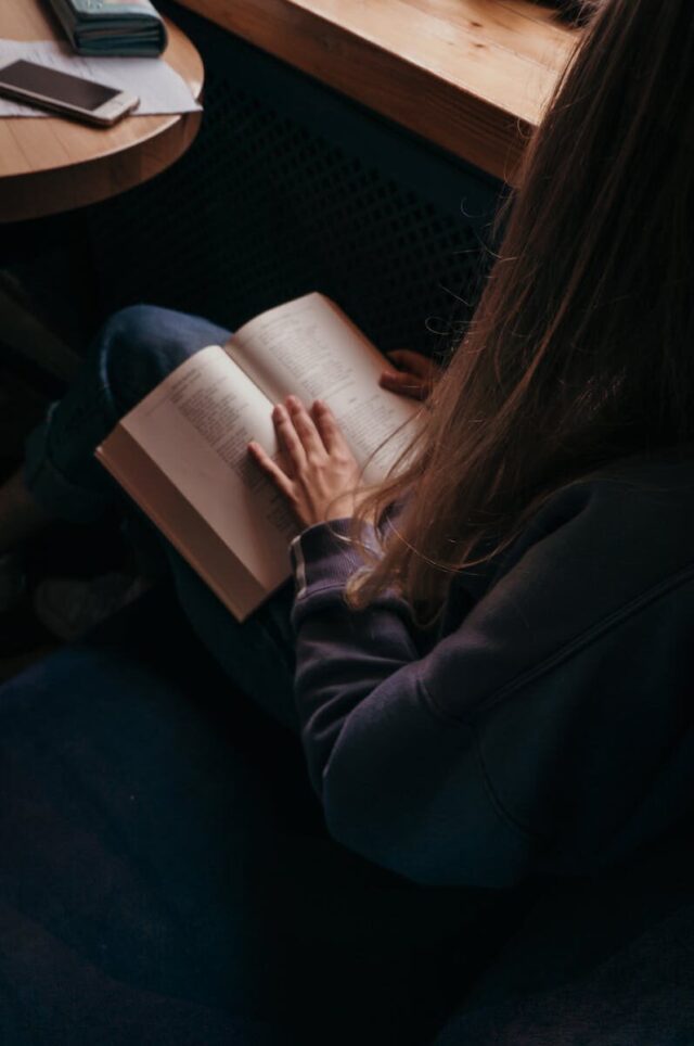 A young woman reading a book in a cozy indoor setting by the window.
