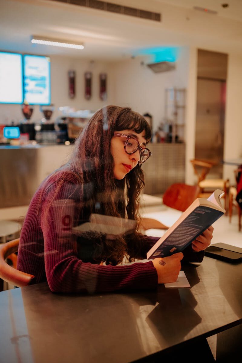 A young woman in a cozy café, absorbed in a book by a window. Captures a moment of relaxation and focus.