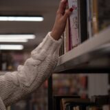 A hand in a cozy sweater reaching for a book on a library shelf, evoking a sense of curiosity and warmth.