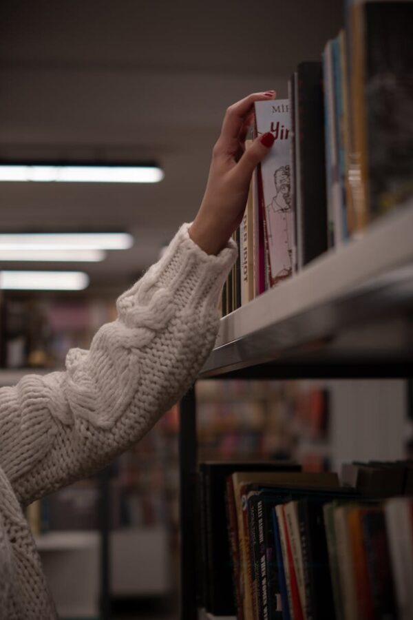 A hand in a cozy sweater reaching for a book on a library shelf, evoking a sense of curiosity and warmth.