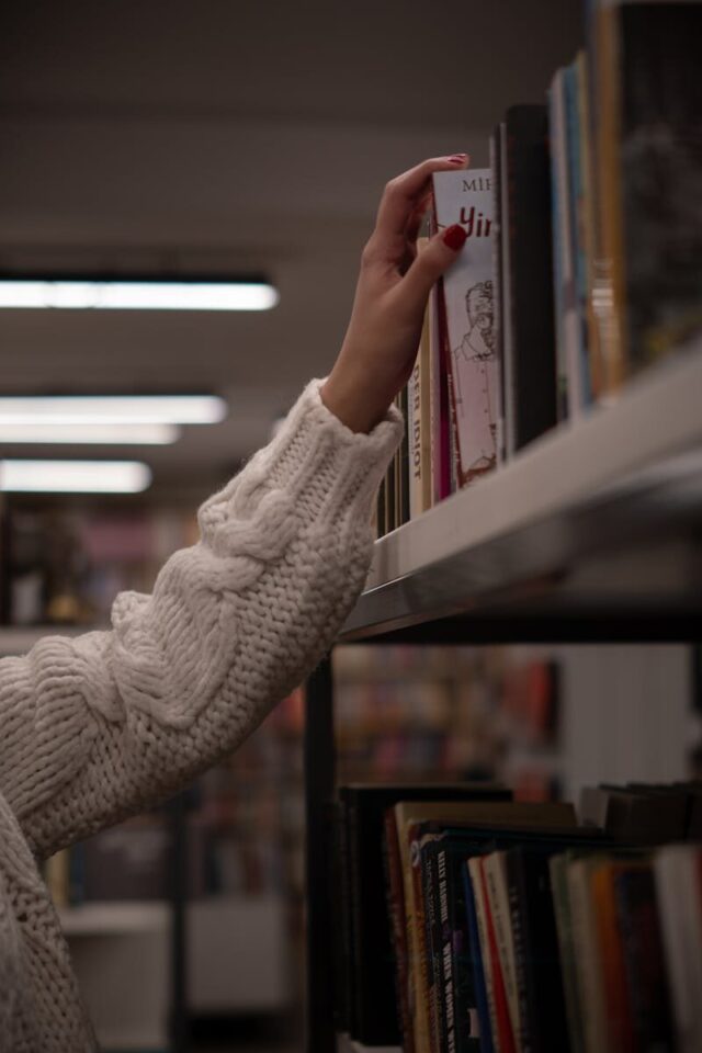 A hand in a cozy sweater reaching for a book on a library shelf, evoking a sense of curiosity and warmth.