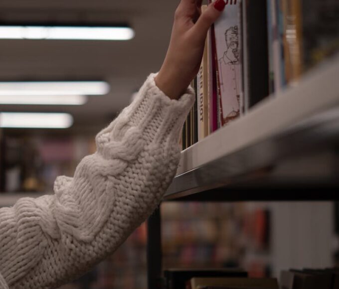 A hand in a cozy sweater reaching for a book on a library shelf, evoking a sense of curiosity and warmth.