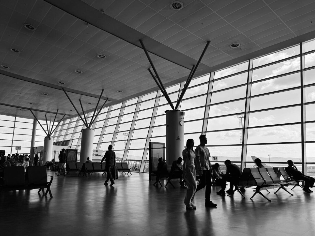 Black and white view of a busy airport terminal with travelers waiting and walking.