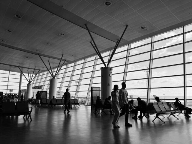 Black and white view of a busy airport terminal with travelers waiting and walking.