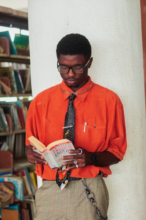 A stylish man in an orange shirt reads a French-English dictionary in a library.