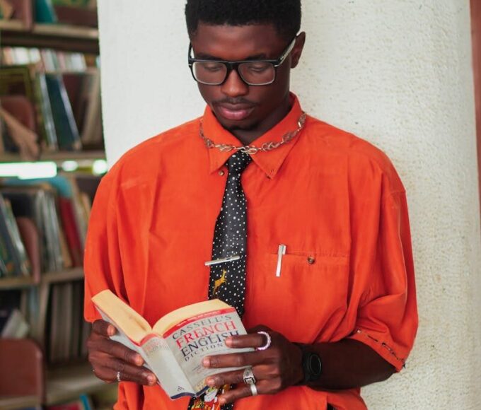 A stylish man in an orange shirt reads a French-English dictionary in a library.