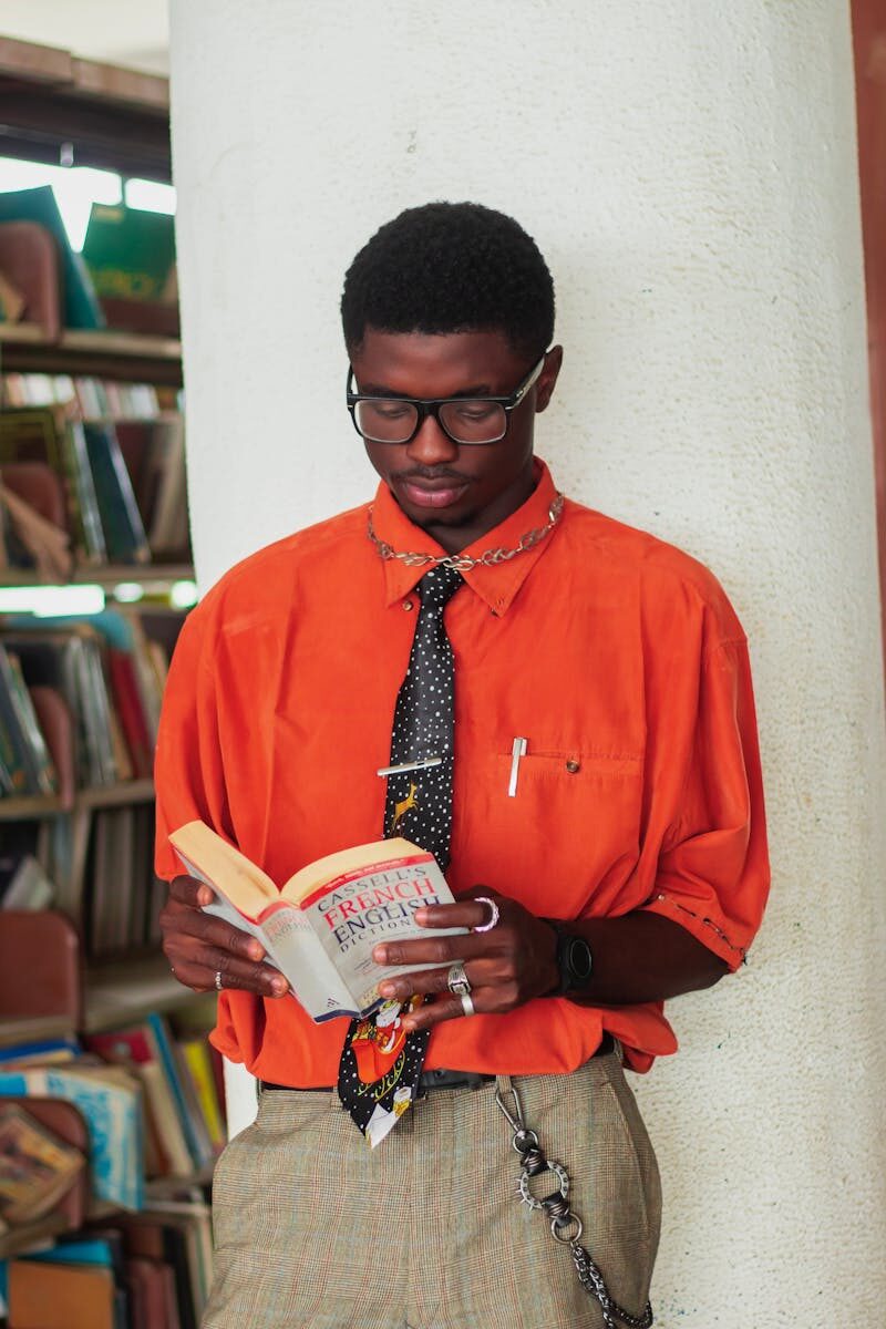 A stylish man in an orange shirt reads a French-English dictionary in a library.