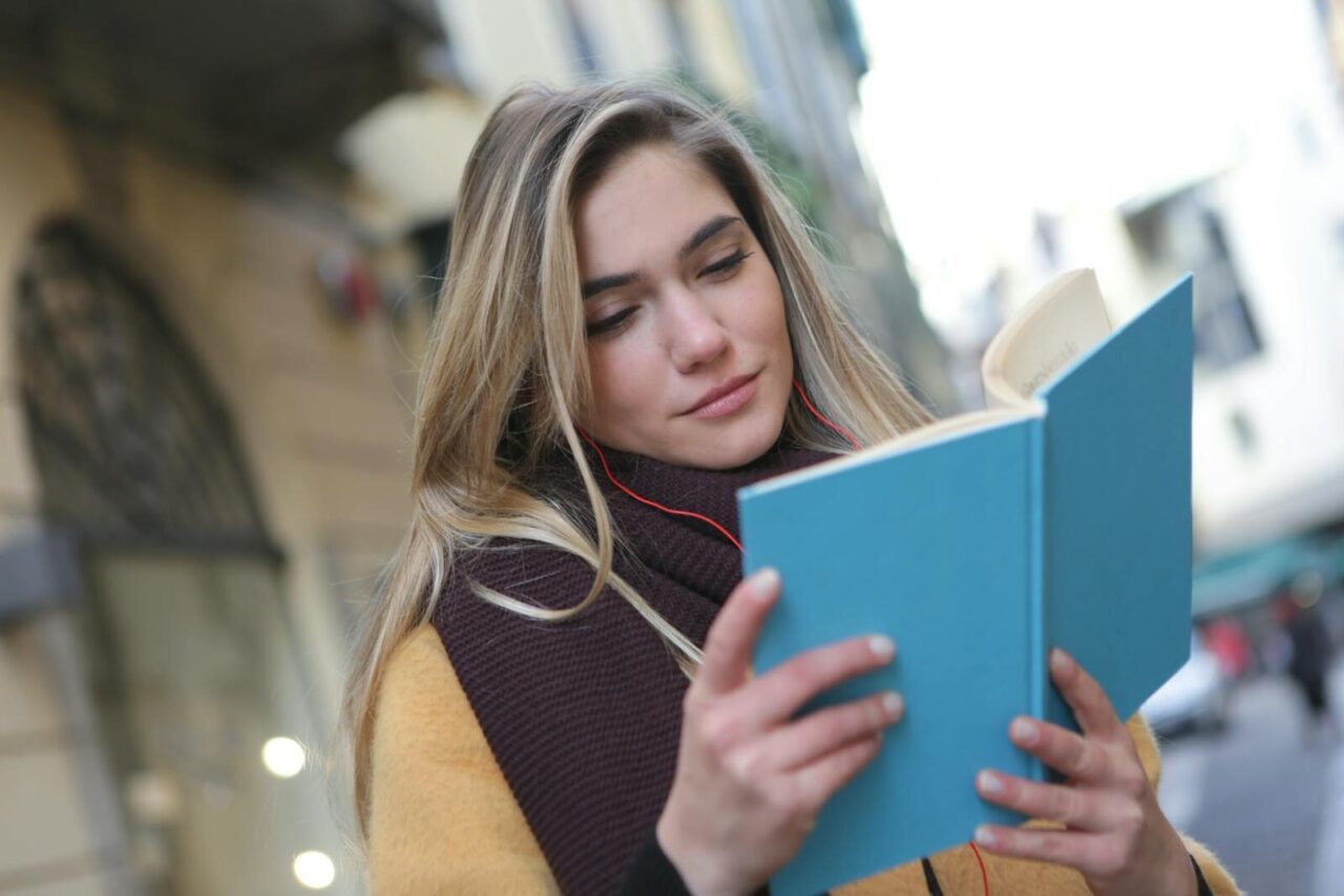 A young woman enjoys reading a book while listening to music on a city street.
