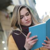 A young woman enjoys reading a book while listening to music on a city street.