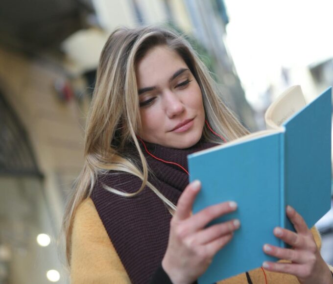 A young woman enjoys reading a book while listening to music on a city street.