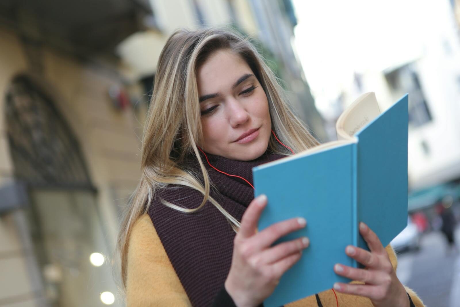 A young woman enjoys reading a book while listening to music on a city street.