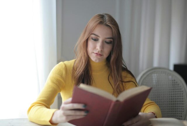 A young woman in a yellow sweater reads a book indoors, enjoying a calm and leisurely moment.