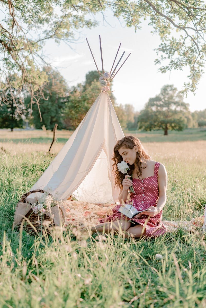 A woman in a polka dot dress enjoys a book and flower in a tranquil park setting with a tent.