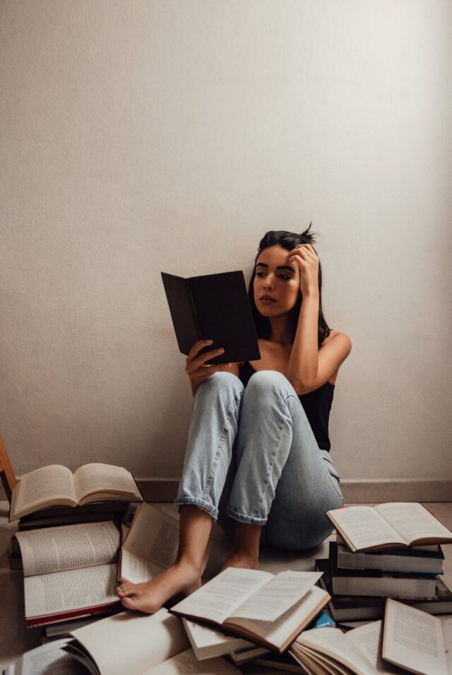 A young woman sitting indoors, deeply focused on reading a book with open books scattered around.