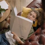 A young woman is lying on a sofa reading a book in a cozy indoor setting.