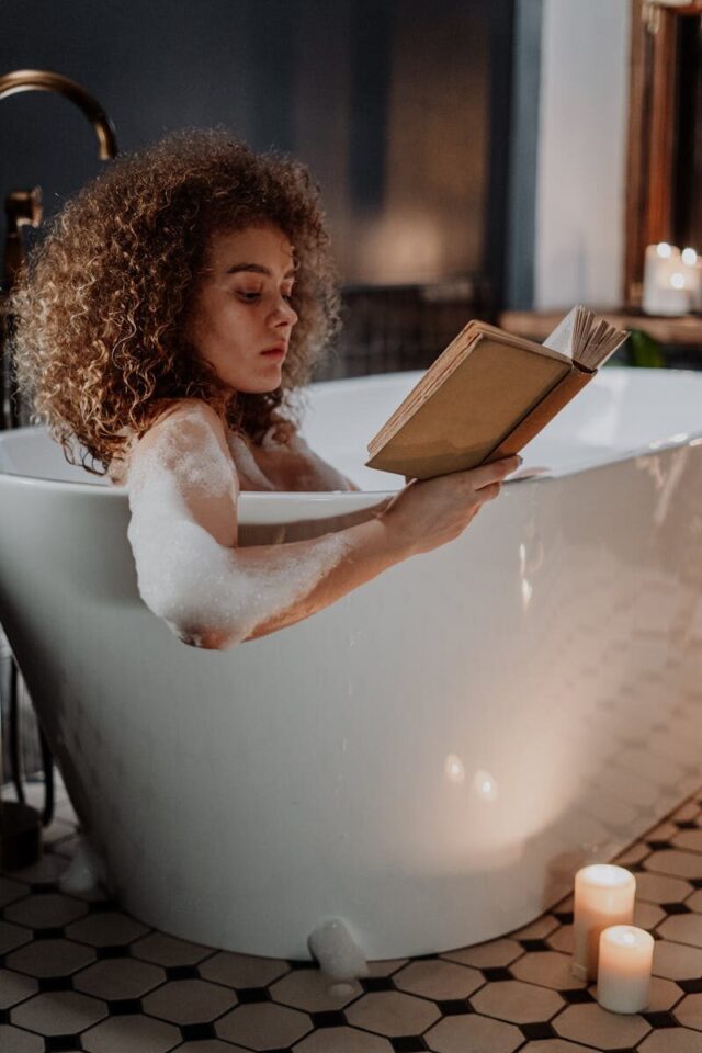 A woman relaxes in a bubble bath reading a book by candlelight, creating a calming atmosphere.