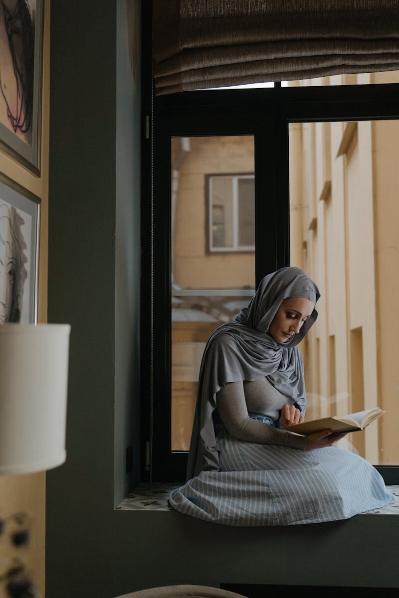 A woman in hijab peacefully reading a book on a comfortable window seat indoors.